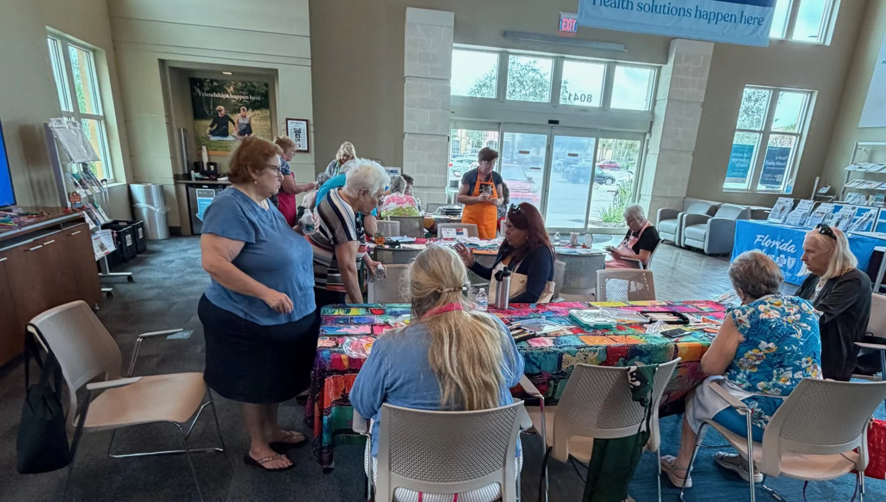 Group of elderly women and one younger woman gathered around a colorful tablecloth at a community event inside a brightly lit room with large windows.