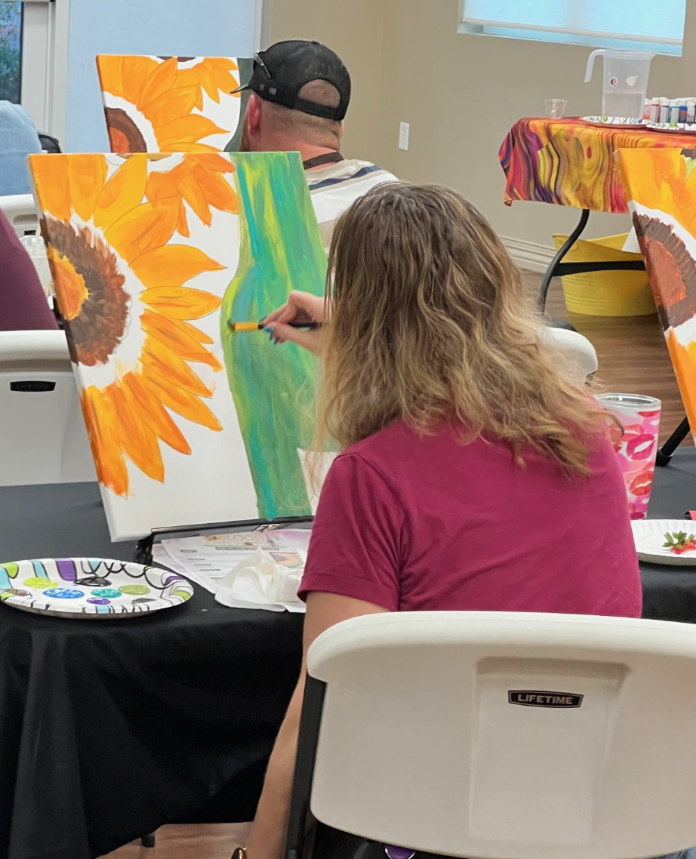 A woman in a maroon shirt painting a canvas with a sunflower and a green vase during a group painting class.