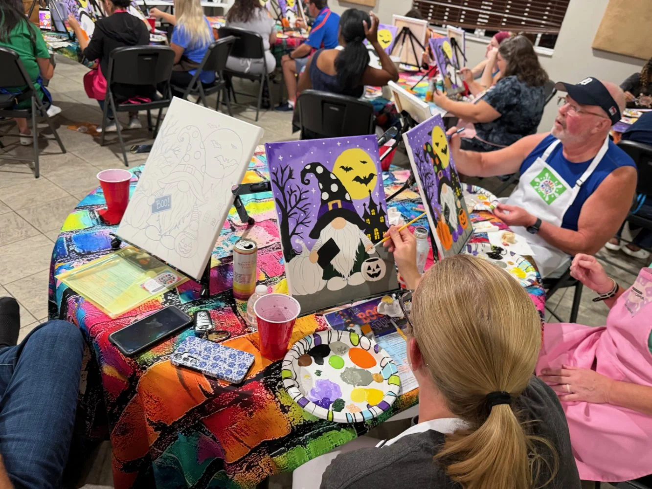People painting Halloween-themed artwork at a group art event, with canvases depicting witches, pumpkins, and spooky scenes.