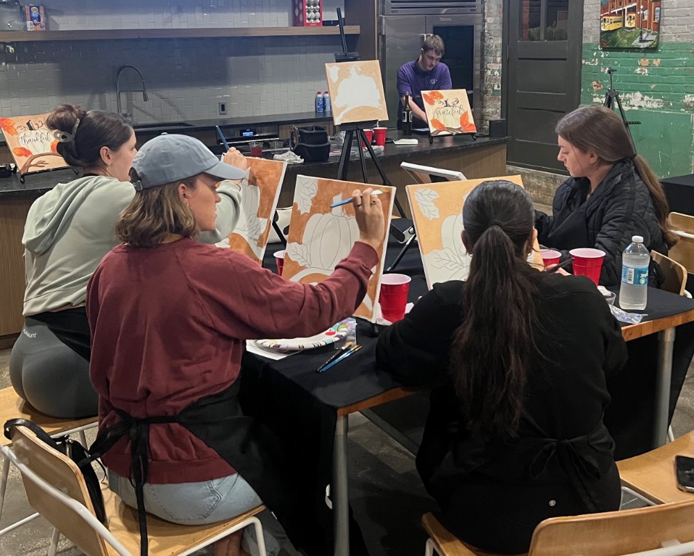 Group of people painting autumn-themed canvases with pumpkins and leaves in an art class