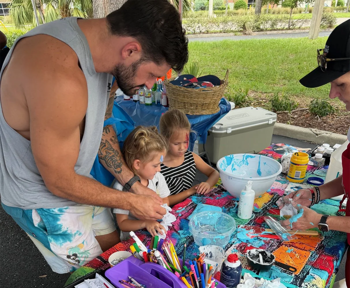 Adults and children decorating cookies at a table covered with colorful cloth, supplies, and icing, outdoors.