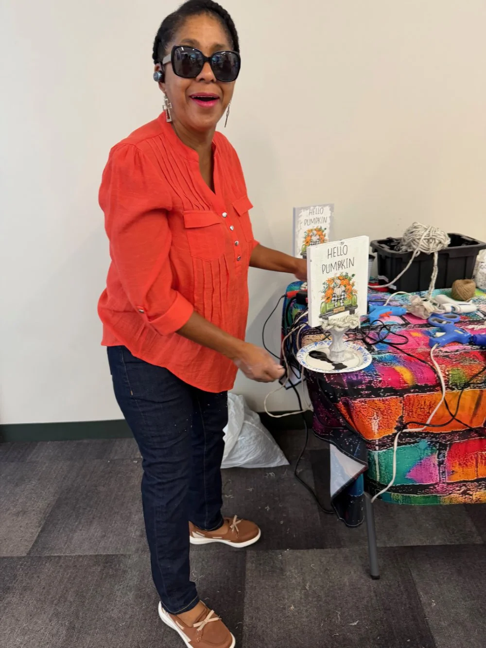 A woman wearing sunglasses in an orange shirt and dark jeans standing at a table with colorful fabric, crafting supplies, and signs that read "Hello Pumpkin."