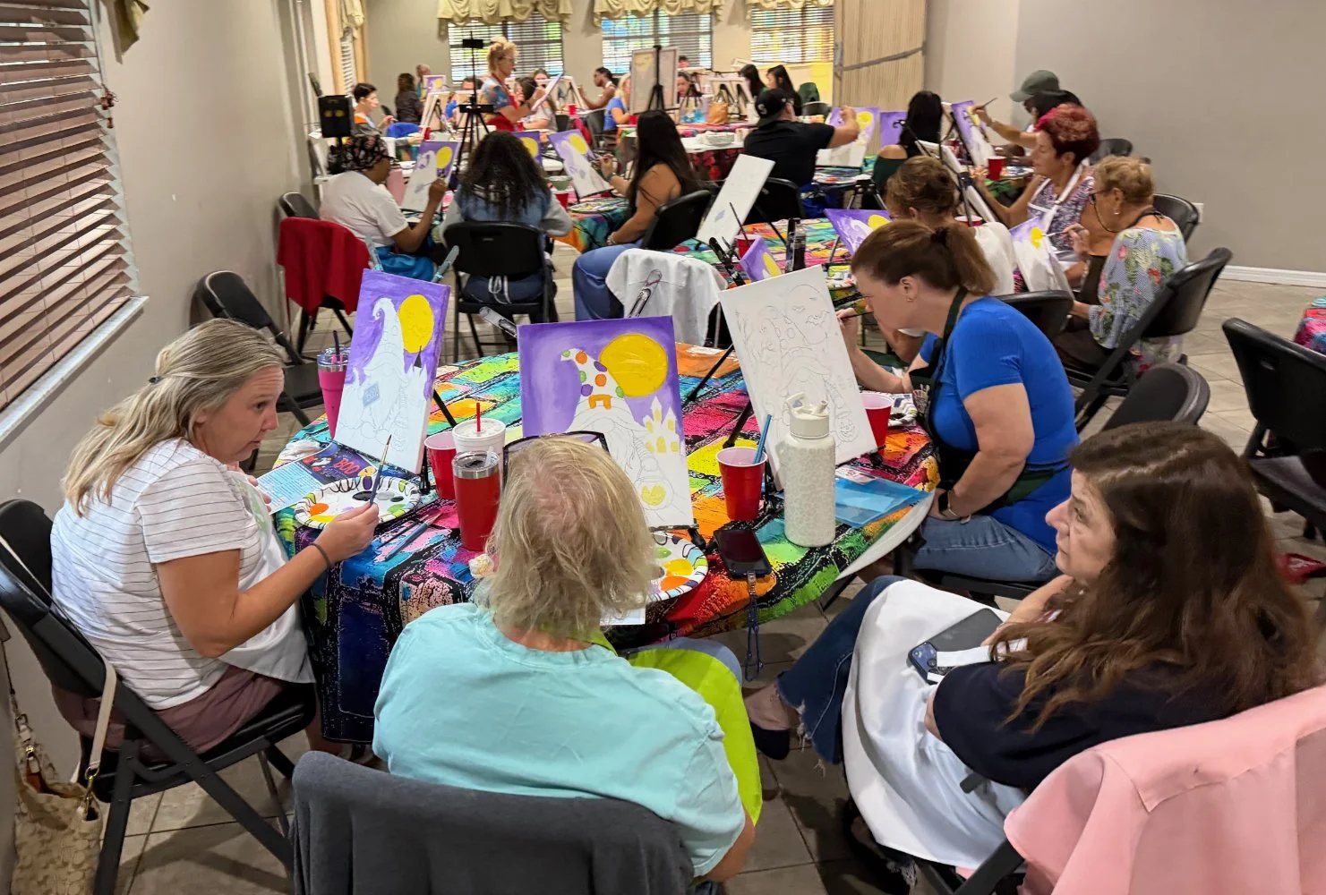 Group of women participating in a painting class, seated around tables with canvases, paint supplies, and drinks, with colorful paintings of unicorns and a moon on display.
