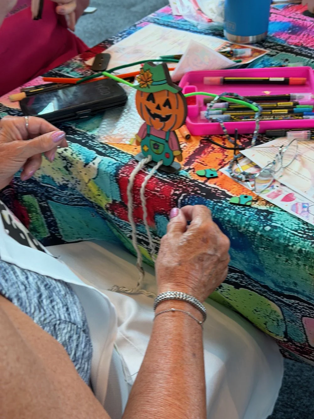Handmade pumpkin decoration with a smiling face, wearing a green hat, and a colorful overall. The table has craft supplies including pens, paper, scissors, and glasses, with a vibrant tablecloth.
