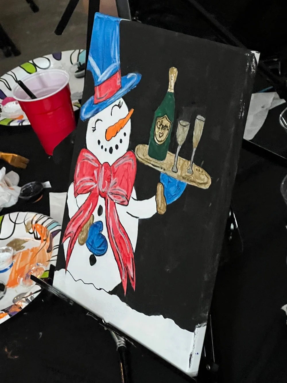 Painting of a snowman dressed as Uncle Sam, holding a tray with champagne and glasses, displayed on an easel surrounded by party supplies.