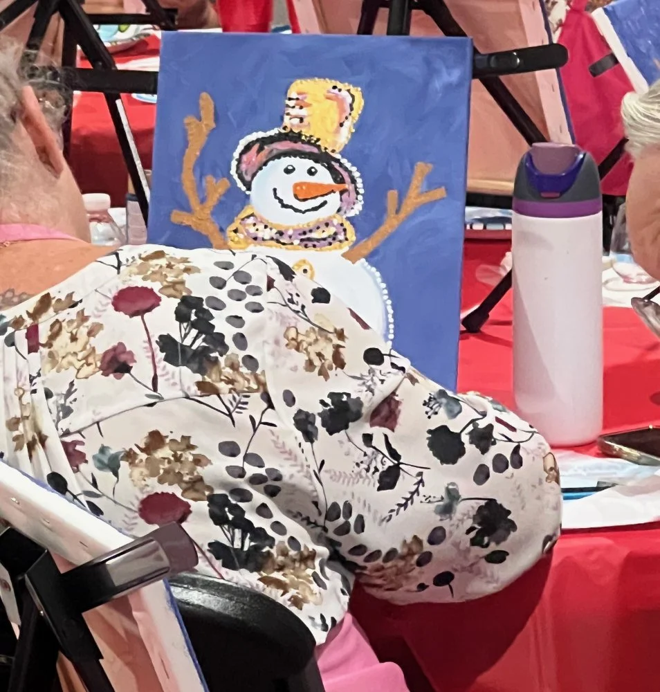 Painting of a snowman with a black top hat, orange nose, and a beaded necklace, set on a blue background at a painting event.