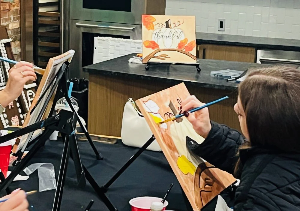 Individuals painting fall-themed canvases with the word 'thankful' outdoors or in a rustic setting.