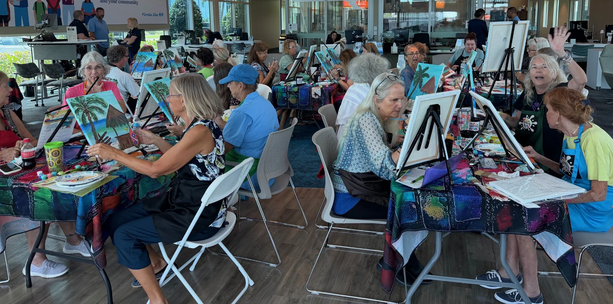Group of older women and a few men participating in a painting class or workshop inside a modern, brightly lit space with large windows, painting on canvases depicting tropical scenes with palm trees and mountains, seated at tables with art supplies 