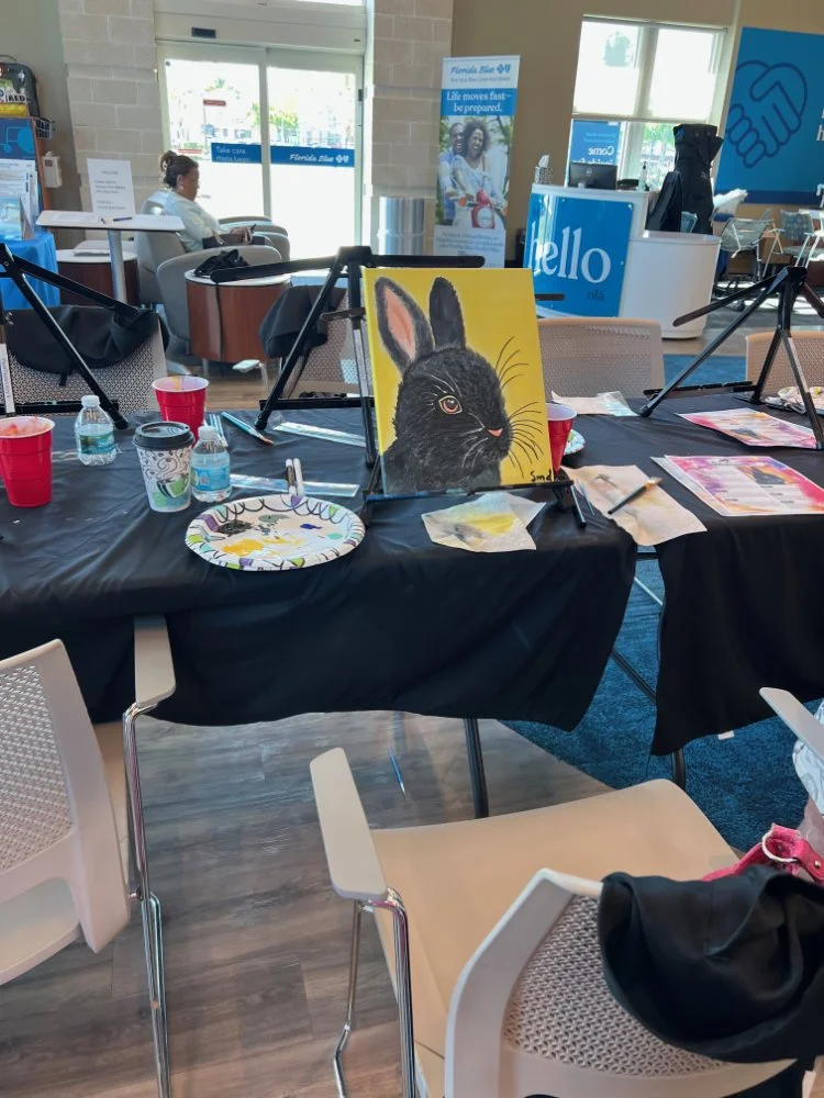 A painting of a black rabbit with pink ears and orange eyes on an easel, set on a table in a busy indoor space with chairs, tables, and people in the background.