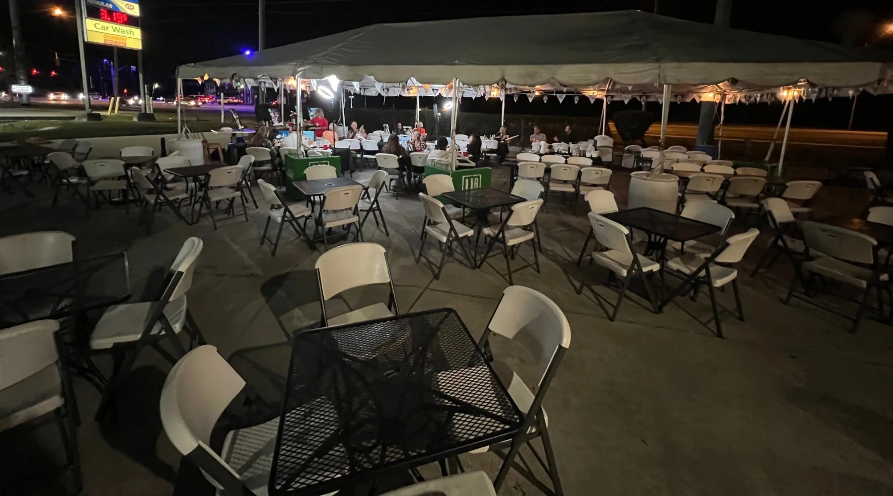 Empty outdoor restaurant seating area at night with white and black tables and chairs under a large canopy, illuminated by string lights, with a car wash sign and some busy street traffic in the background.