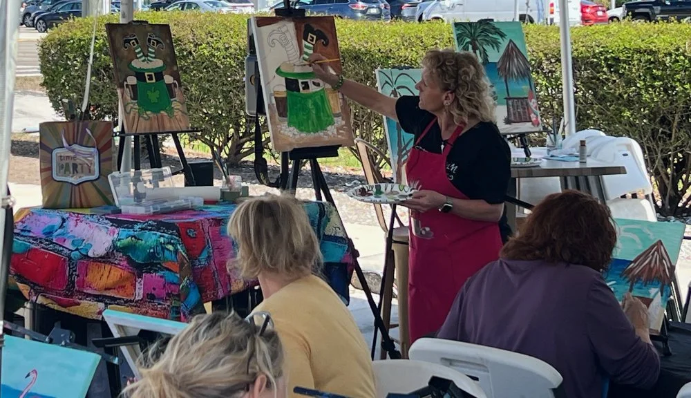 A woman in a black shirt and red apron is painting a canvas at an outdoor art event, with paintings of tropical themes and abstract art displayed around her. People are seated, working on their own artwork.