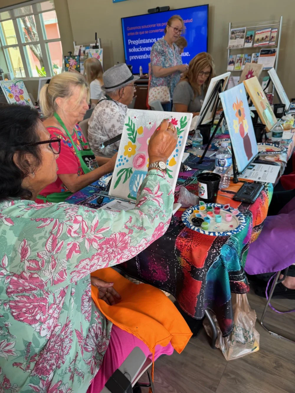 People participating in a painting class with floral artwork, set in a bright room with natural light and colorful decorations.