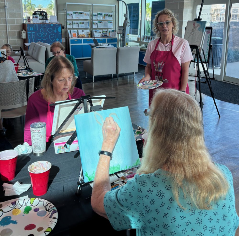 A group of elderly women participating in a painting class at a community center. One woman is painting on a canvas with a light blue sky and green grass, while another woman stands holding a paint palette. Other women are seated at tables with art s
