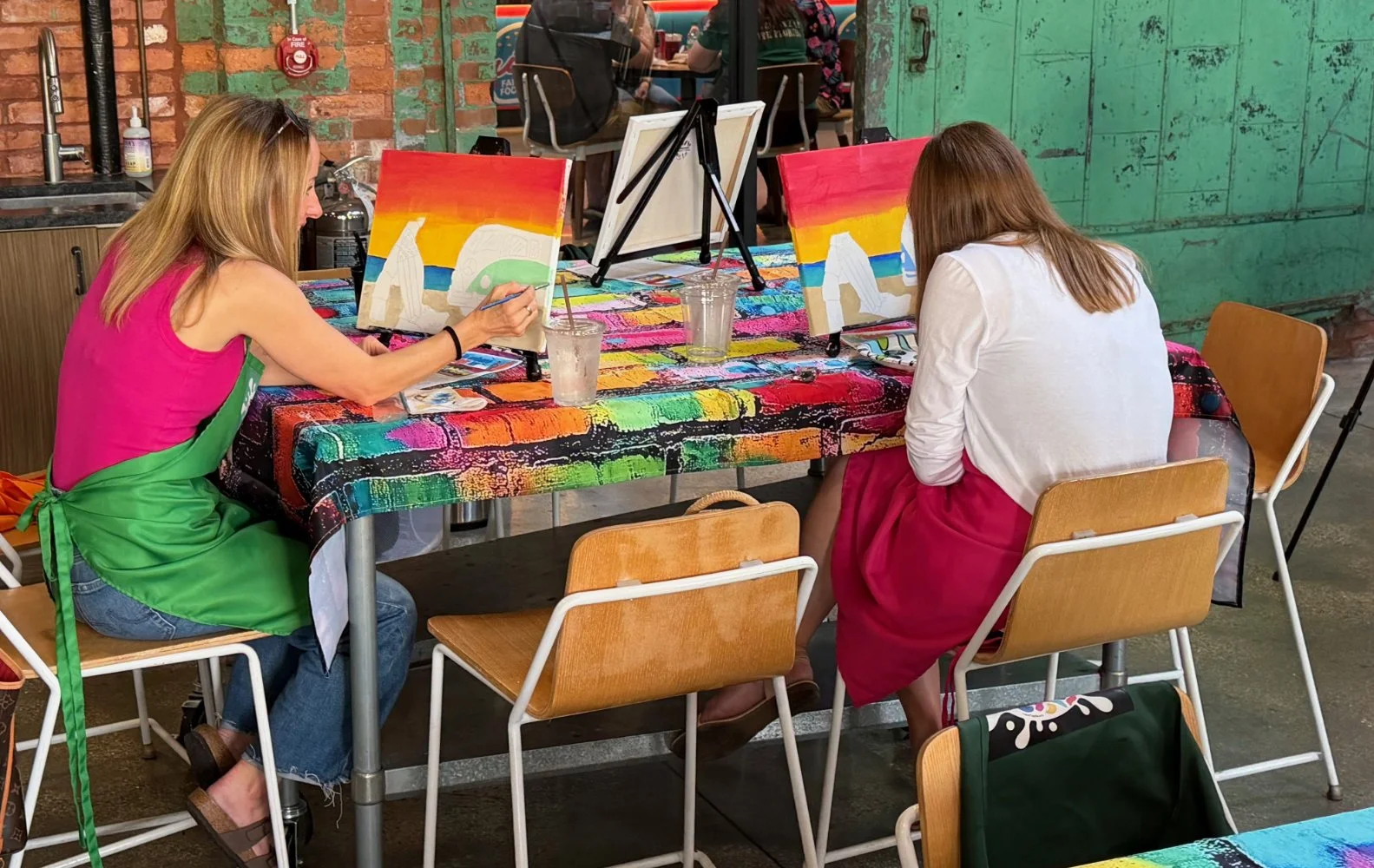 Two women painting on canvases at a table with colorful tablecloths, in a rustic art studio. One woman is wearing a pink top and green apron, the other is in a white top and red skirt. They are focused on their artwork.