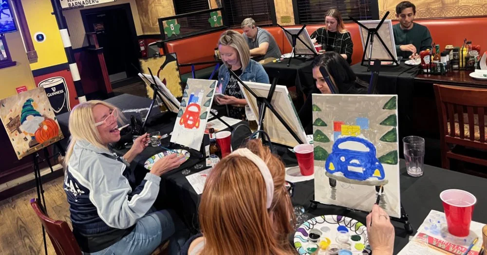 People painting on canvas at a party or class event at a restaurant or bar, with paintings of cars and pumpkins, and smiling woman in foreground.