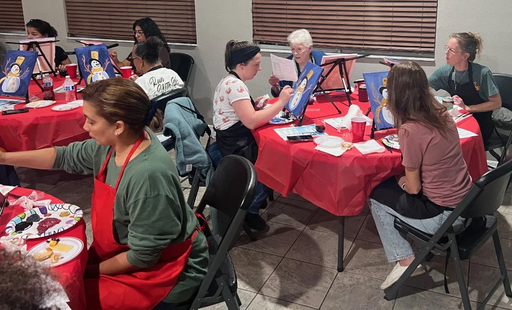 Group of women gathered around tables with red tablecloths, painting snowmen on canvases during a holiday-themed art activity.