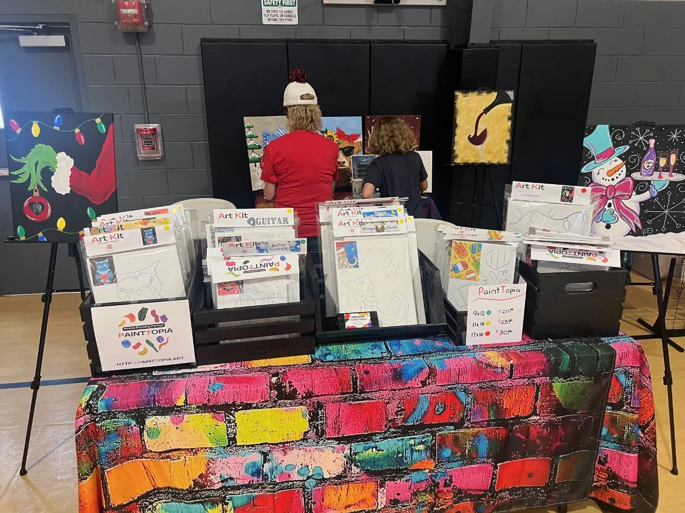 A table with art kits for painting and coloring, featuring Halloween and holiday-themed artwork. Two people, one adult and one child, are painting on canvases in the background. The table is covered with a colorful, graffiti-style tablecloth, and var
