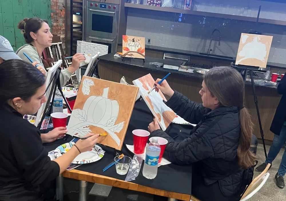 People painting autumn-themed canvases with pumpkins and leaves in a creative workshop.