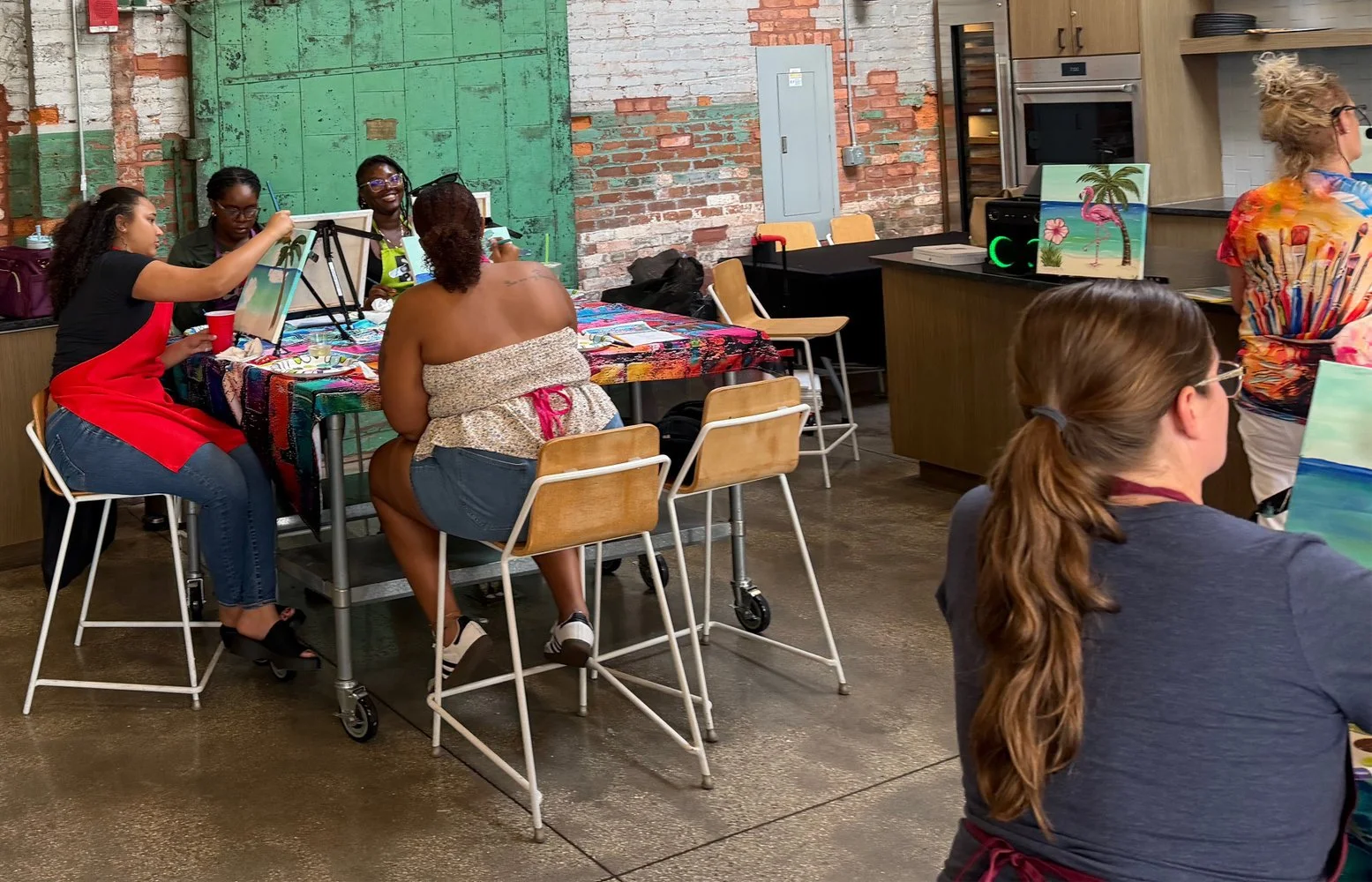 People participating in a painting class indoors with brick wall and kitchen in the background, one woman is painting a tropical scene with pink flamingos on an easel.
