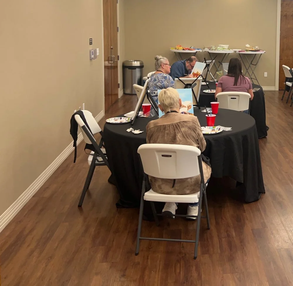 People painting at tables in a room with beige walls and wooden floors, with plates and cups on the tables and snacks on a tray in the background.