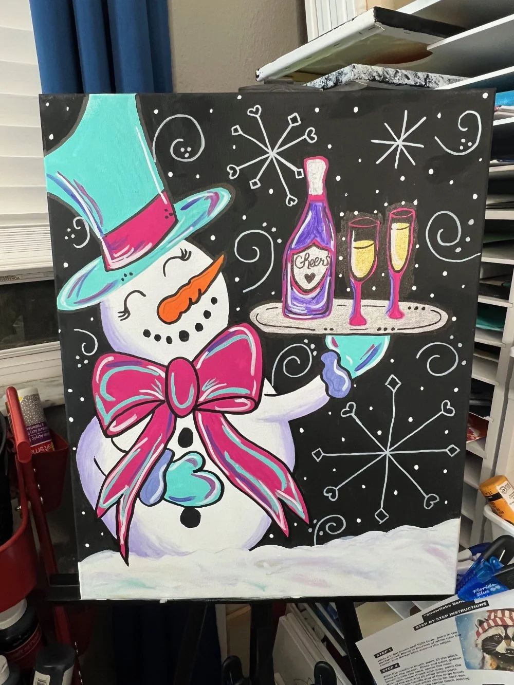 A colorful snowman wearing a purple top hat with pink accents, a pink bow tie, and decorated with a heart-shaped button. The snowman is holding a tray with a bottle labeled 'Cheers' and two glasses of champagne. The background is black with white sno