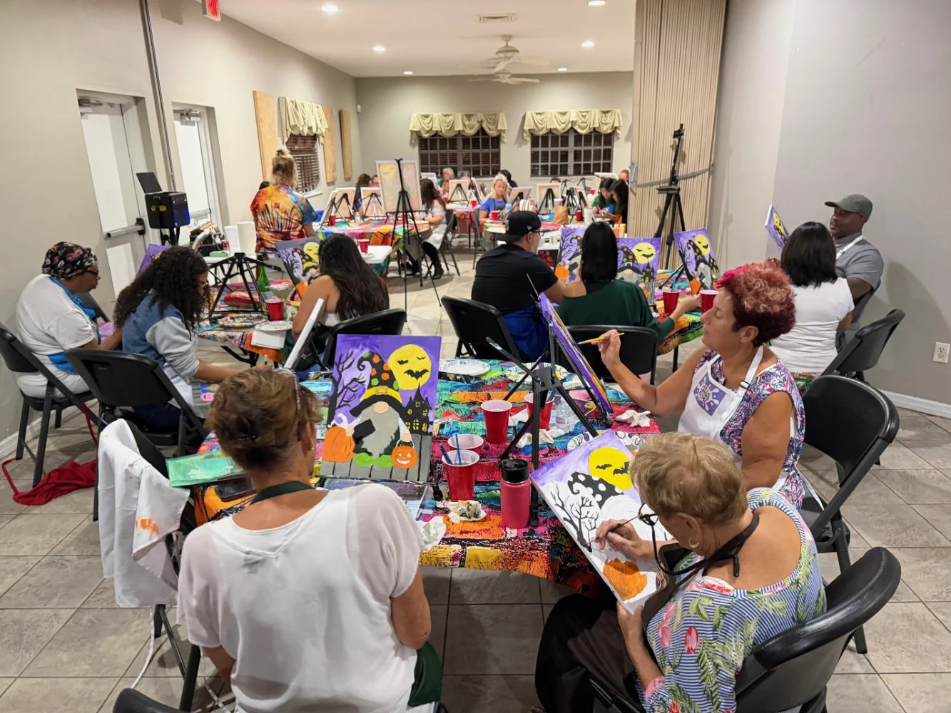 People at a Halloween-themed painting class, sitting at tables and painting spooky Halloween scenes on canvases with colorful backgrounds, in a room decorated with yellow valances and balloons.