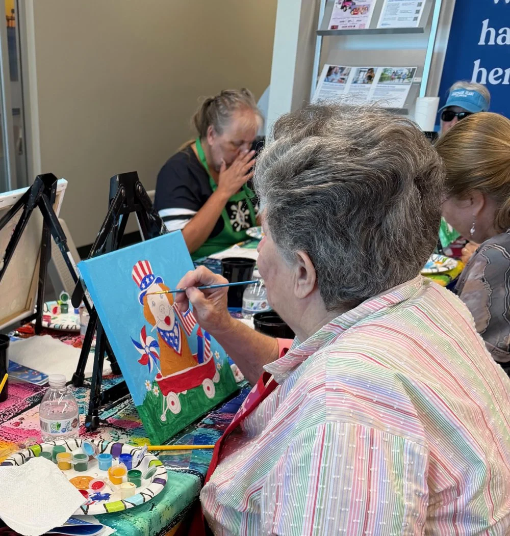 An elderly woman is painting a patriotic picture of a dog in a red wagon as part of an art event. She is using a small paintbrush and has a palette of paint in front of her. Several other people are seated around the table, some with arts and crafts 