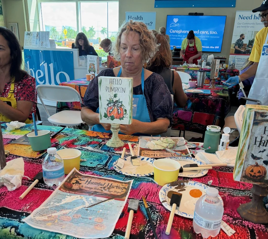 Woman at craft table holding a painted sign that says 'Hello Pumpkin' with a pumpkin illustration. Other people are seated and working on craft projects at tables covered with colorful tablecloths, craft supplies, paper, and paint. Large windows in t