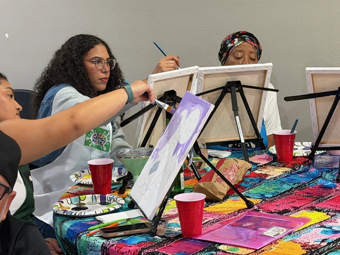 People participating in a painting class, sitting at a colorful table with easels, paint supplies, and cups.