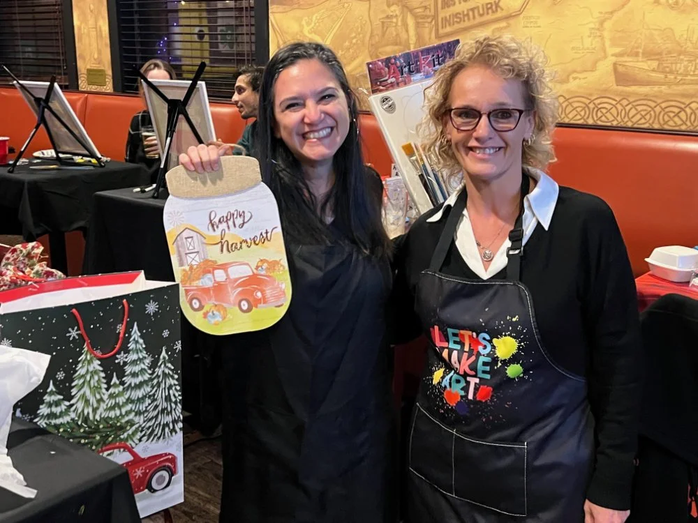 Two women smiling at a craft event, one holding a harvest-themed cutting board with a red truck illustration, surrounded by gift bags and painting supplies.