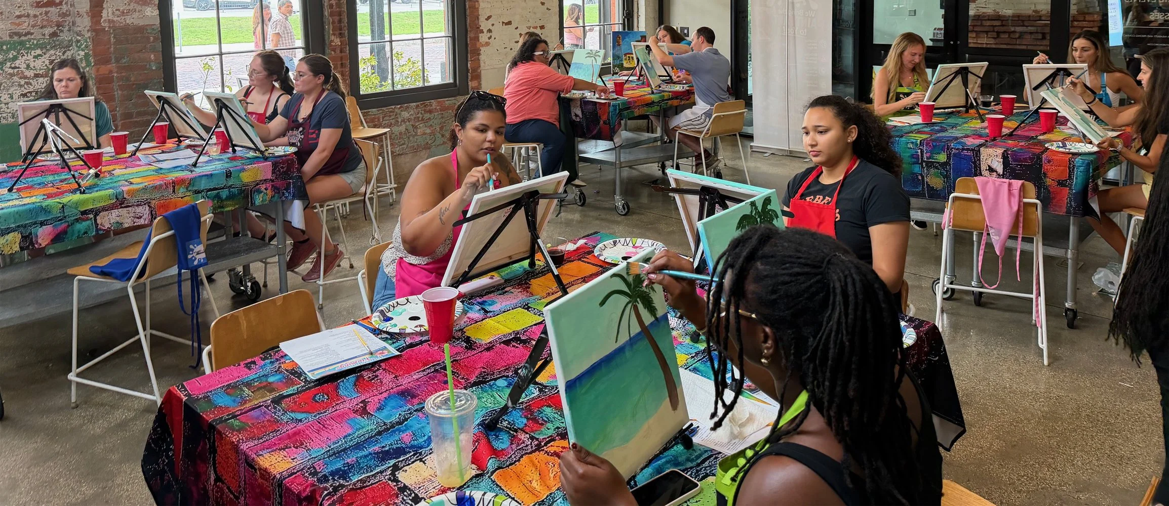 A group of women participating in a painting class in a gallery-style space. They are working on paintings of tropical scenes, with colorful tables covered in vibrant, abstract tablecloths. The room has large windows, exposed brick walls, and appears