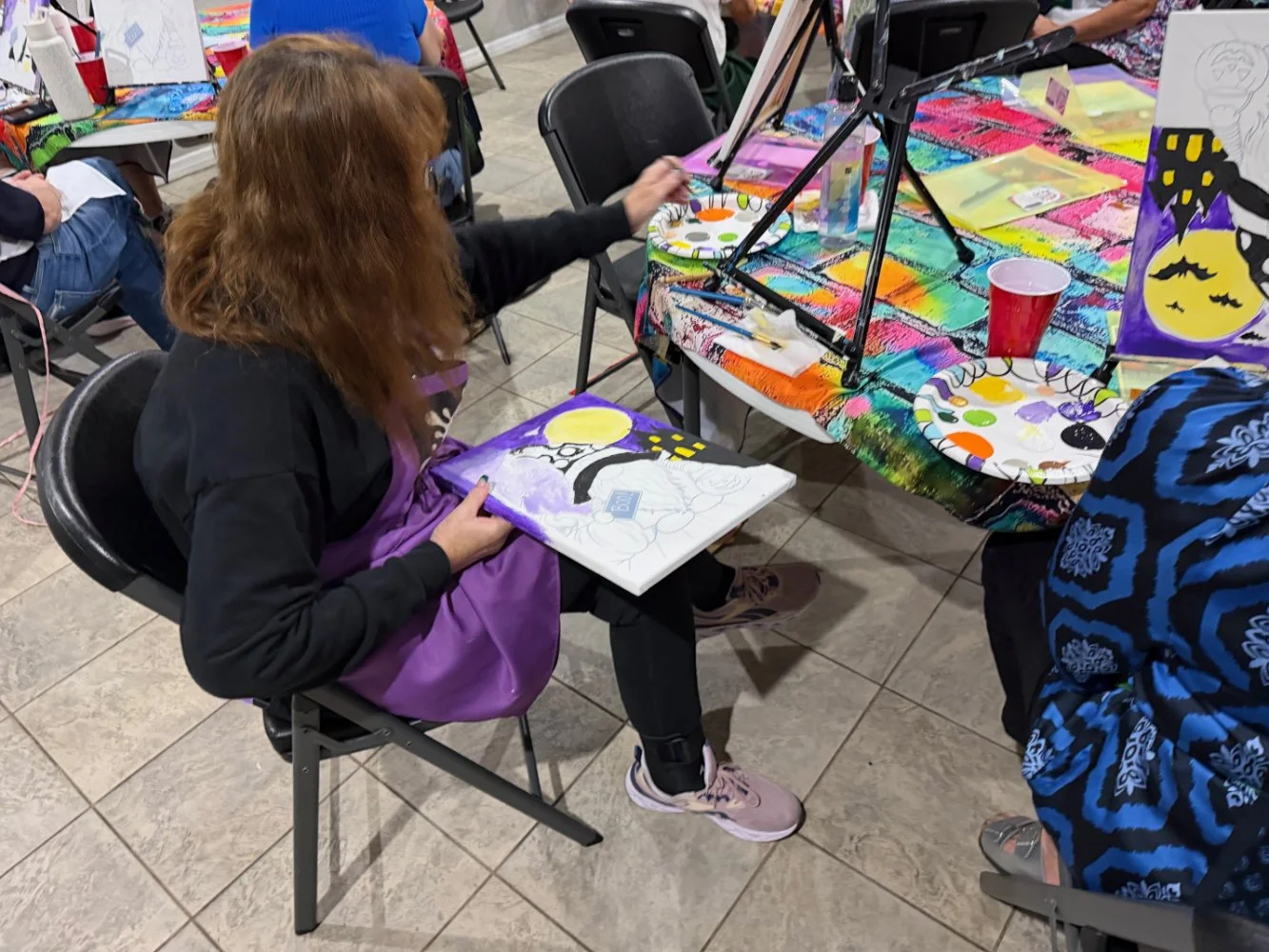 A woman with red hair sitting on a black chair, painting a picture with Halloween-themed motifs such as a yellow moon, a black cat, and a purple background, while holding a canvas. The table in front of her has various art supplies, colorful decorati