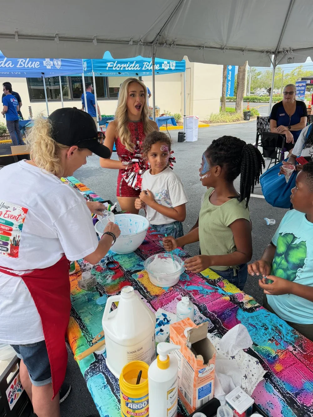 Children and adults at a face painting and craft booth under a tent at an outdoor event, with face paint supplies and a colorful table covering.
