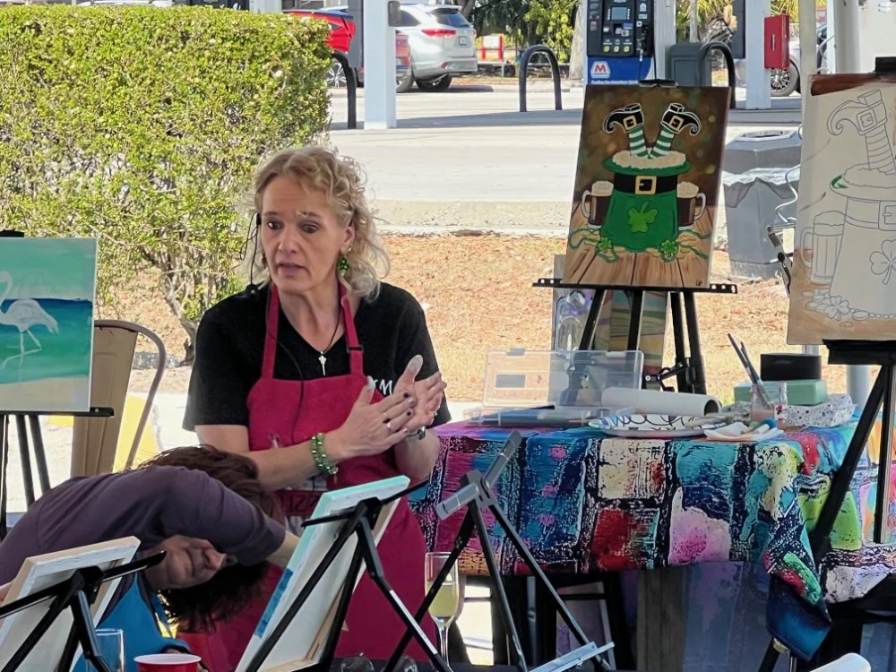 Woman in black shirt and red apron speaking at a painting class, surrounded by paintings, canvases, and art supplies, with a backdrop of parked cars and a hedge outside.