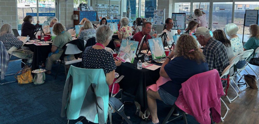 People participating in a painting class at a community center or art studio, sitting at tables with art supplies, creating paintings, with large windows in the background.