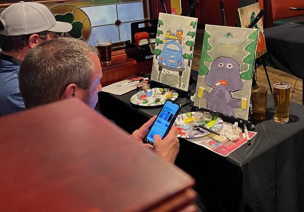 People sitting at a table watching paint-along artwork, with two canvases showing cartoon-like paintings of a car and a purple creature drinking beer, in a restaurant or pub setting.
