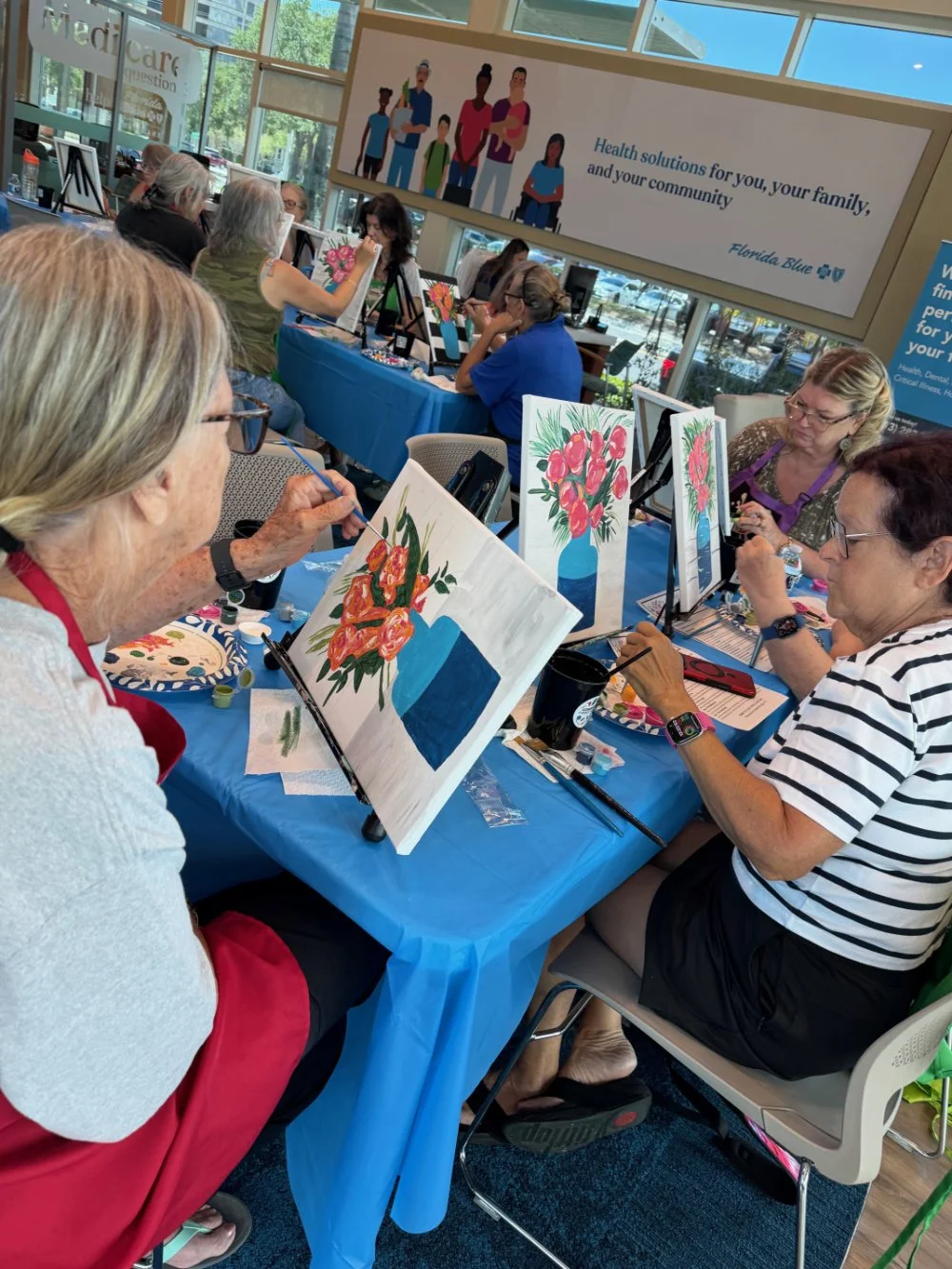Group of women participating in a painting event at a community center, painting floral still life on canvases. Large windows and colorful artwork are visible in the background.
