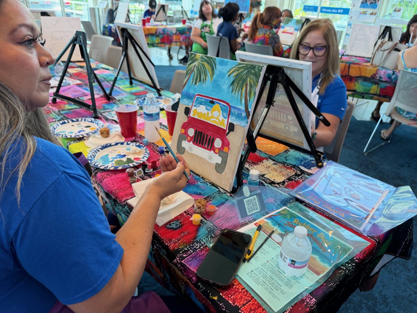 Group of women participating in a painting class, painting beach scenes with palm trees and a red jeep at a colorful table.