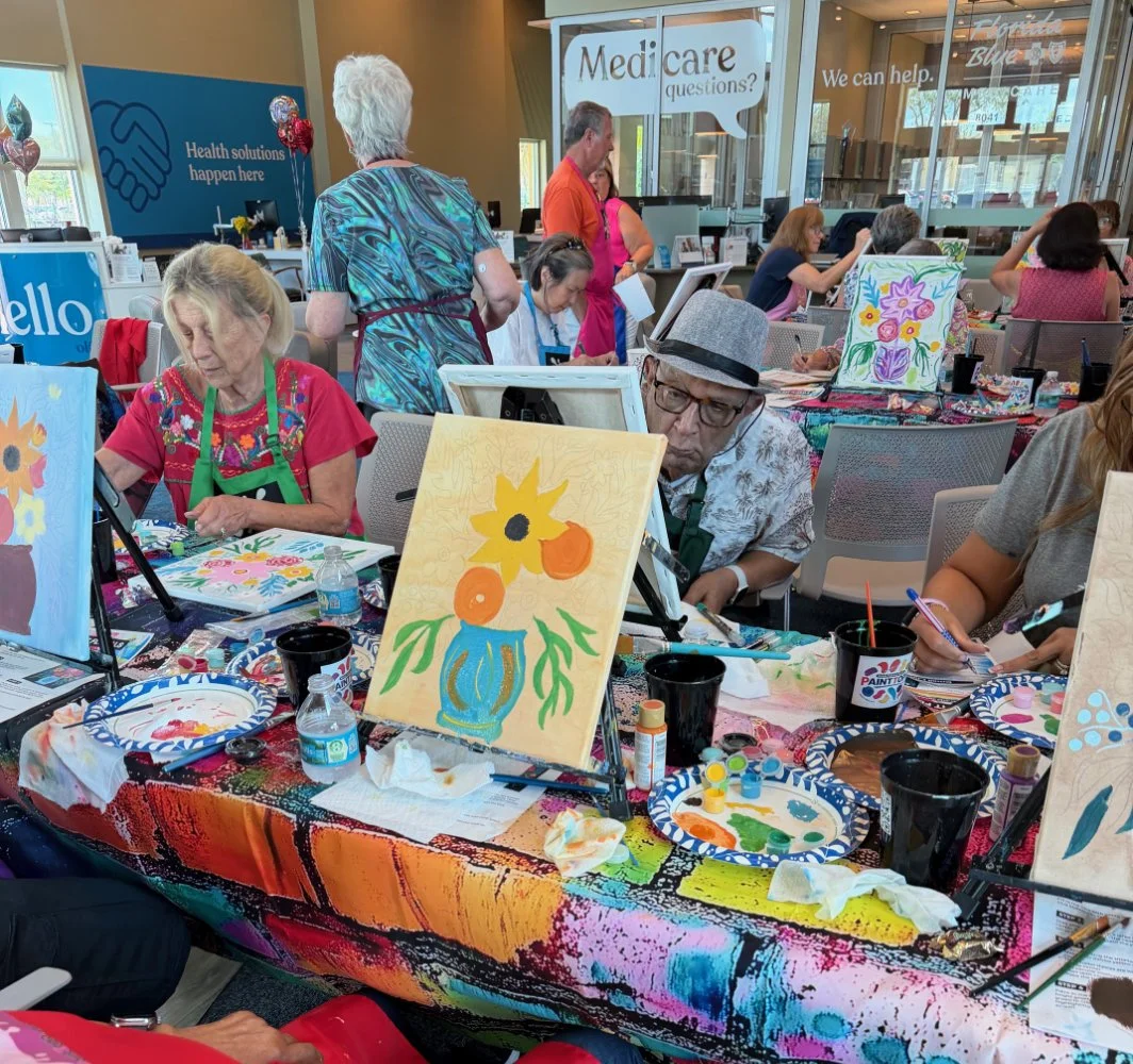 People participating in a painting class in a brightly lit room, with paintings of flowers on canvases and art supplies on tables, including paint, brushes, and cups.