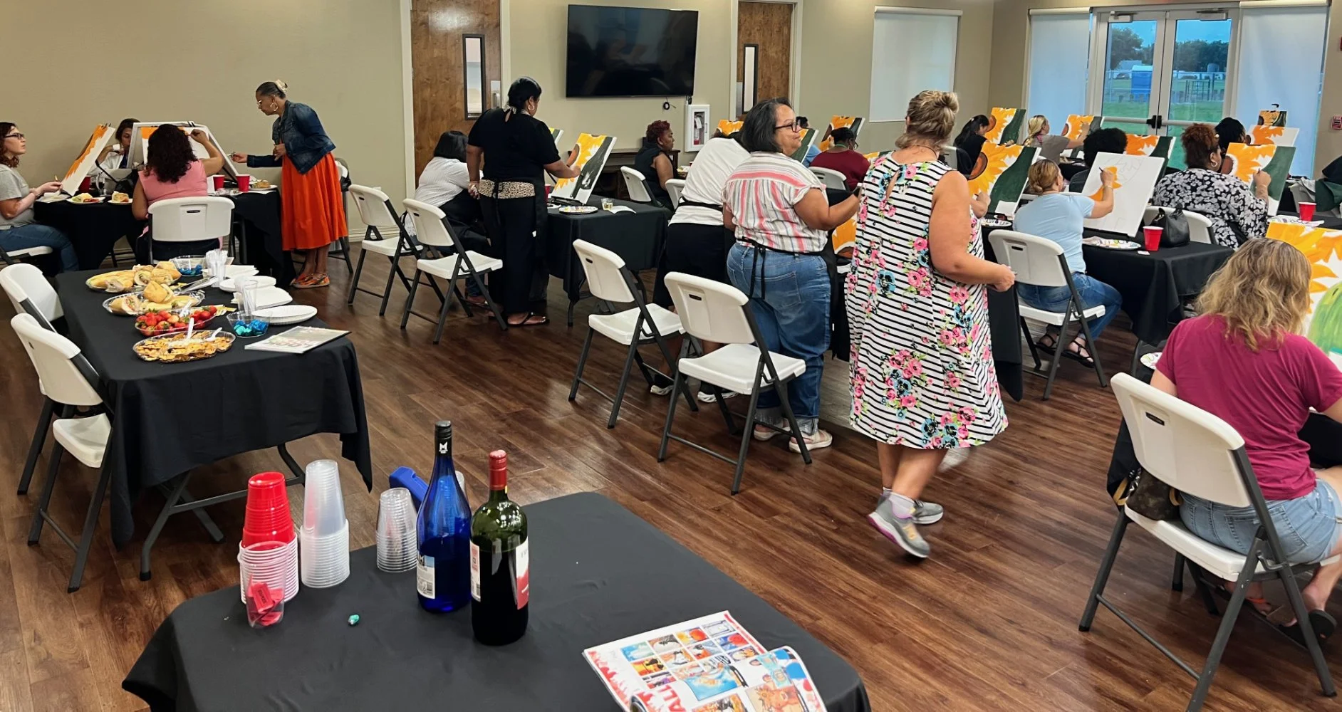 People participating in a group painting activity in a well-lit room with large windows and a TV on the wall. Some are painting at easels, others are walking and socializing, with food and drinks on tables nearby.