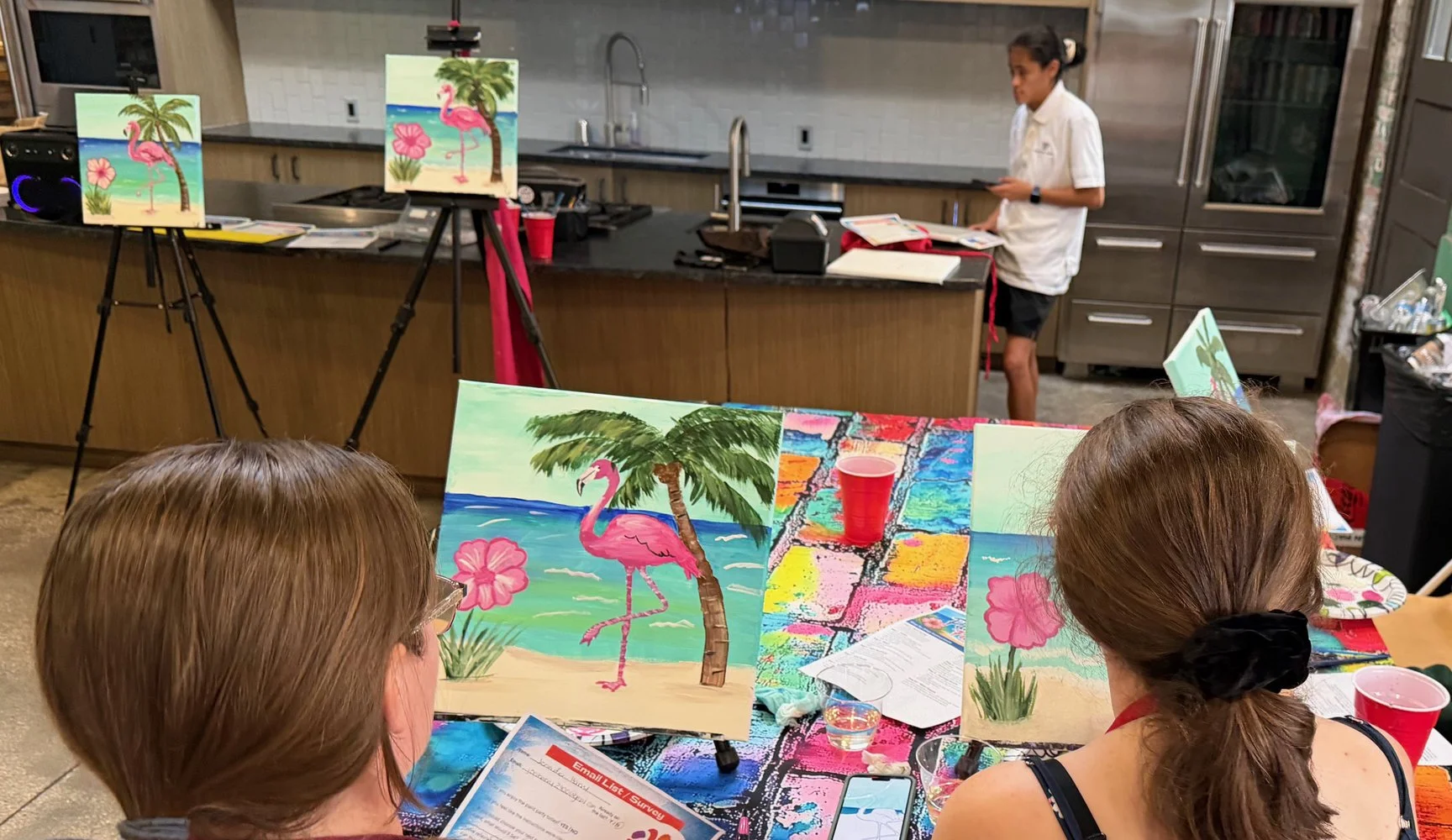 Children participating in a painting class with tropical beach and flamingo themed canvases, in a kitchen or community room setting.