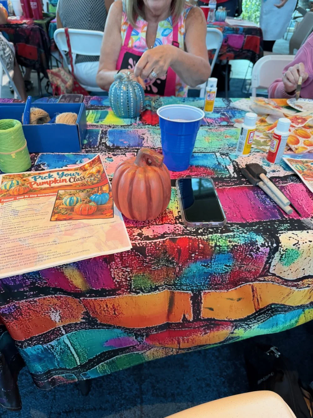 A table with colorful, vibrant tablecloth, decorated with pumpkins, craft supplies, glue, paint, and a pamphlet about pumpkin painting. In the background, a woman is engaged in a craft activity.