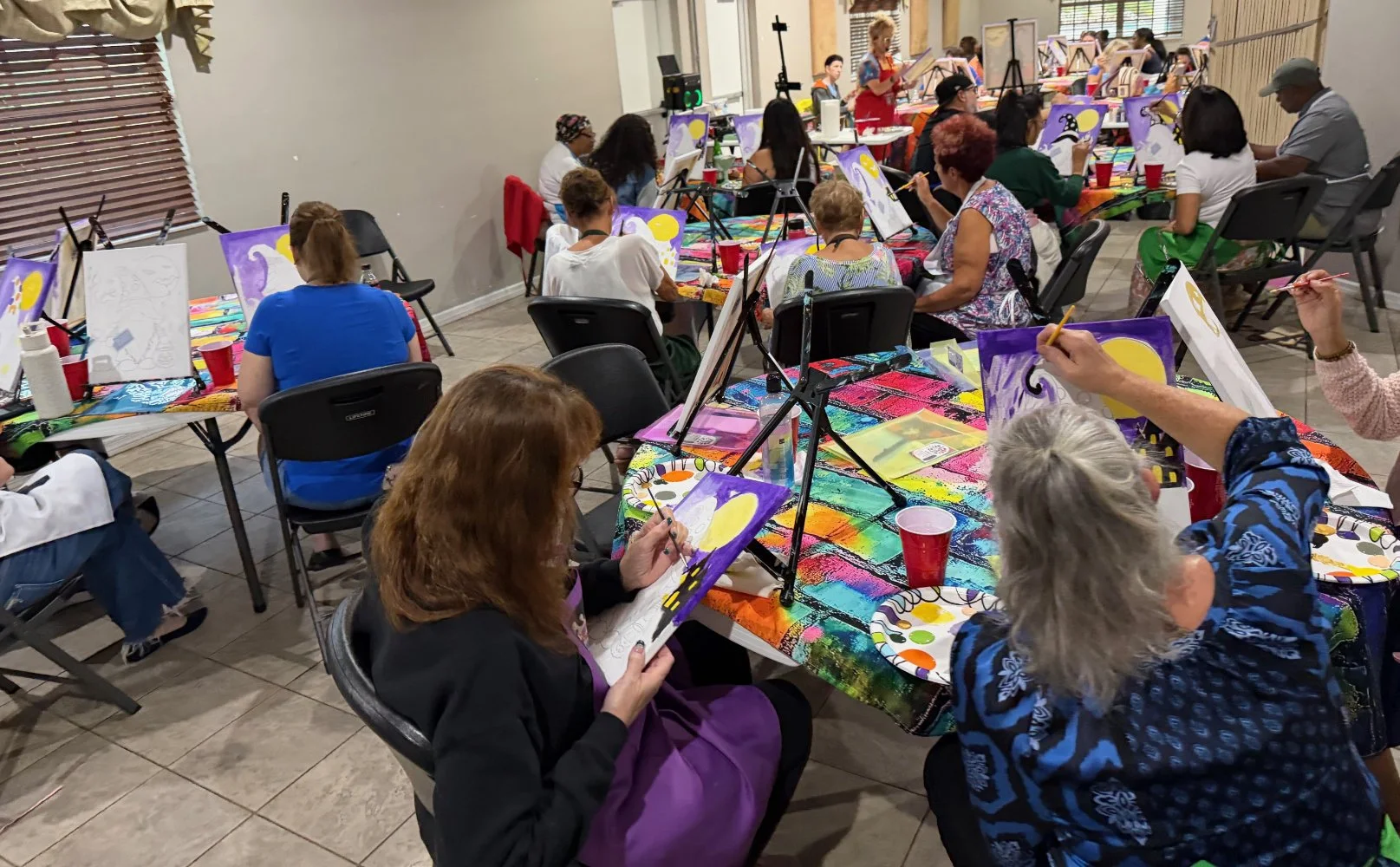 Group of people participating in a paint and sip class, painting colorful scenes of a moon and clouds on canvases in a decorated indoor space.