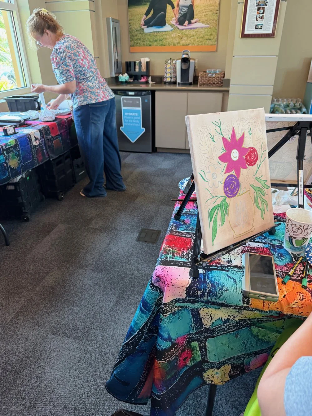 Woman standing at table with art supplies, preparing for a painting activity in a room decorated with colorful art and a floral painting on an easel.