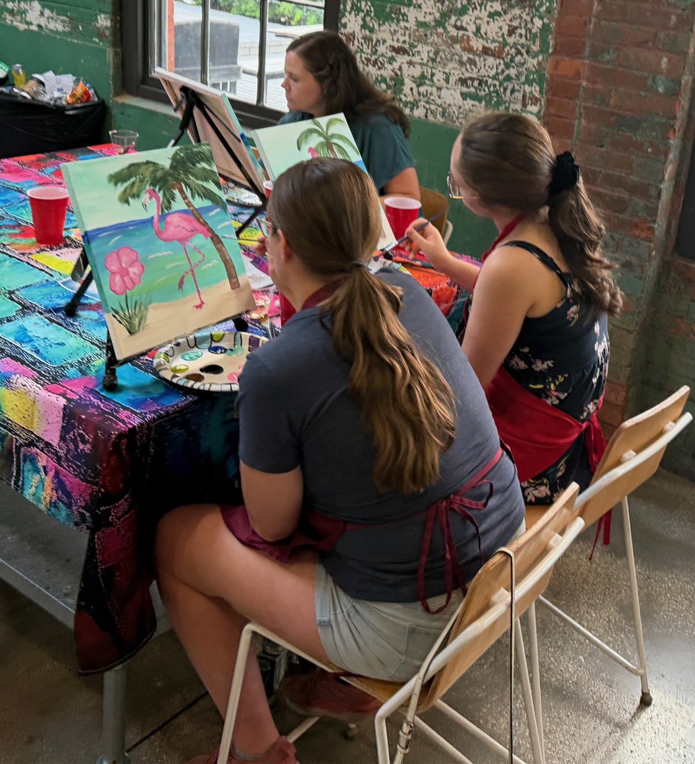 Three women painting tropical scene with a pink flamingo, palm trees, and a flower at an indoor art class.