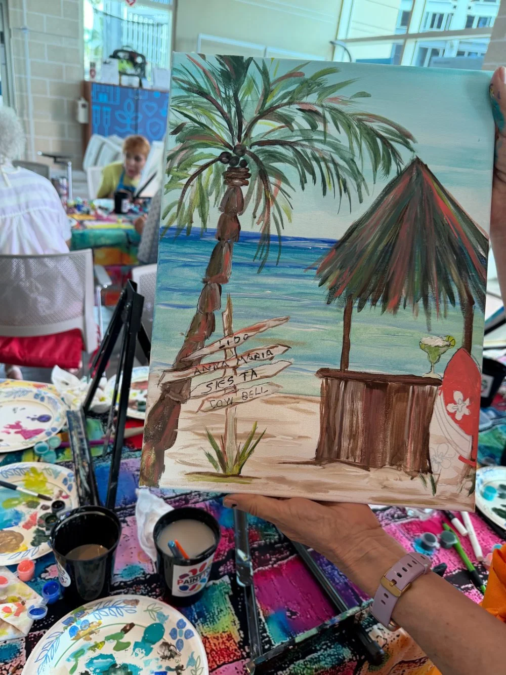 Painting of a tropical beach scene with a palm tree, a thatched hut, and a cocktail, held by a person at an art class, with women sitting at tables in the background.