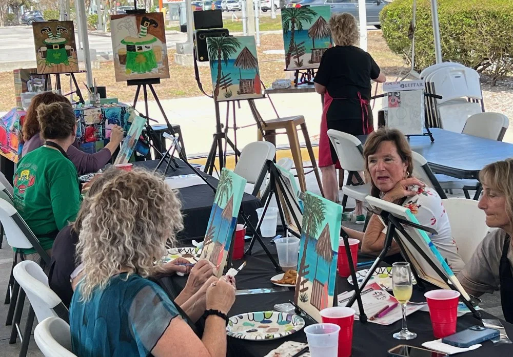 People participating in a group painting class, creating tropical beach scenes and huts outdoors.