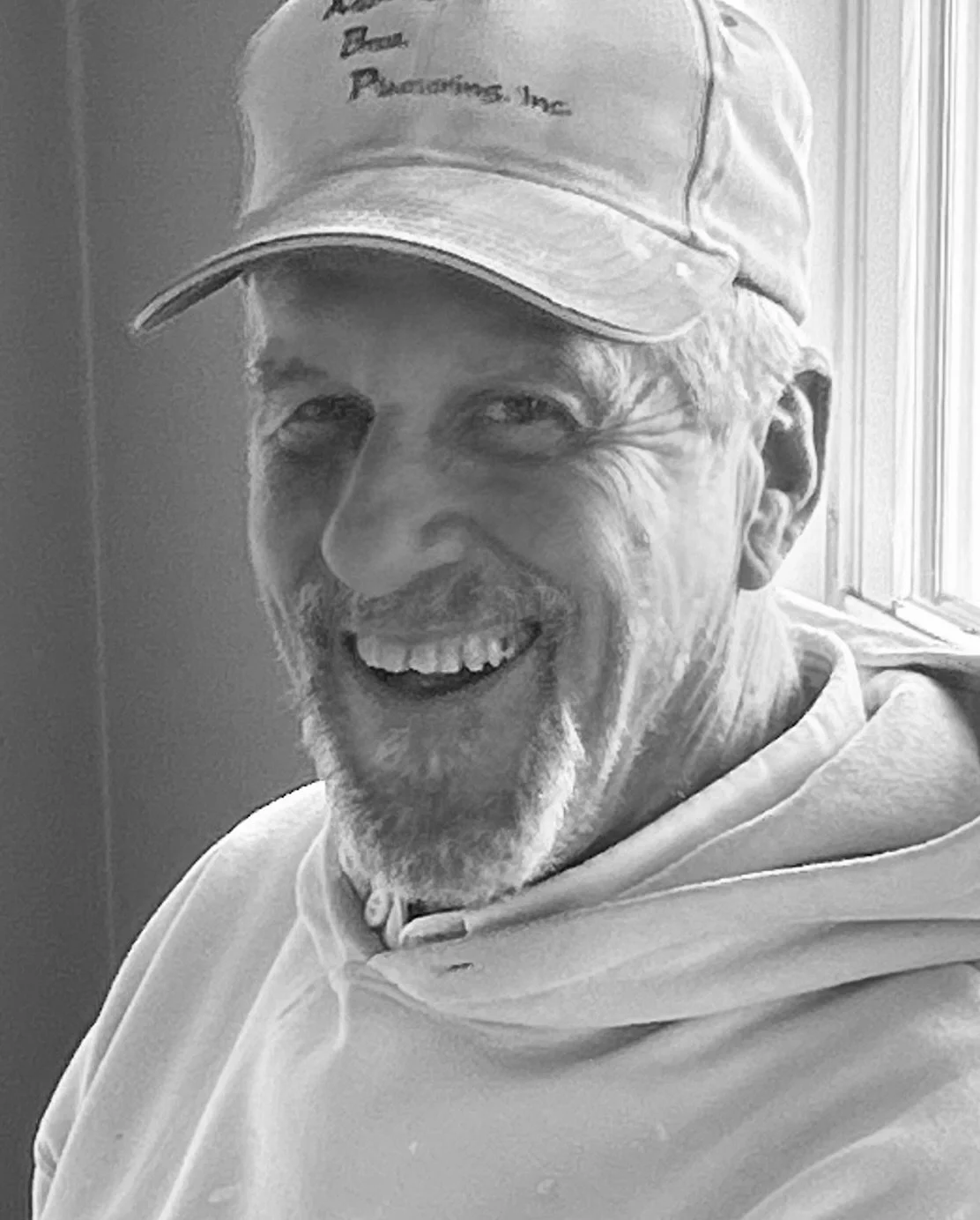 A smiling elderly man wearing a cap and hoodie, sitting near a window.