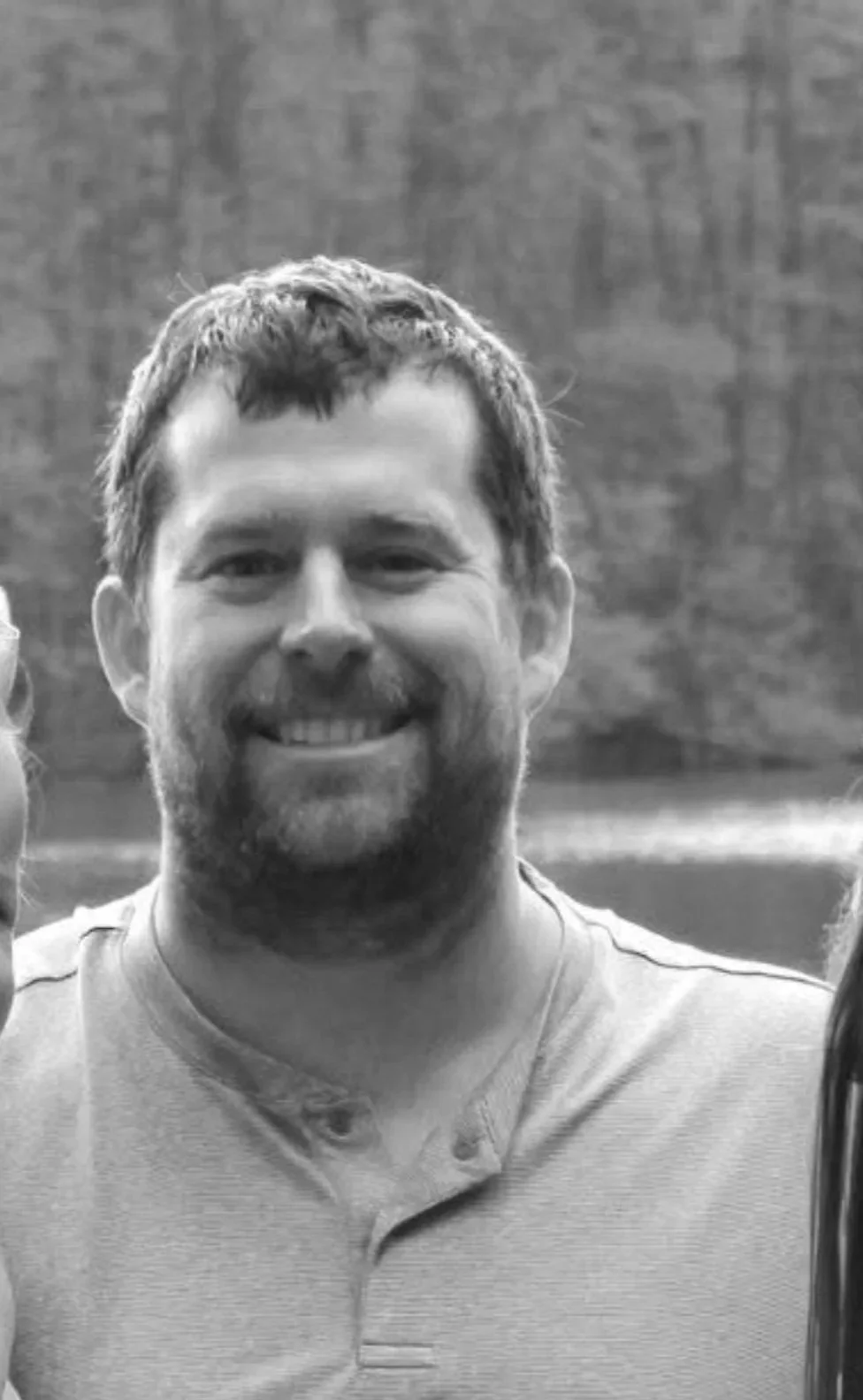 A smiling man with short hair and a beard, wearing a casual shirt, is standing outdoors with a brick wall in the background.