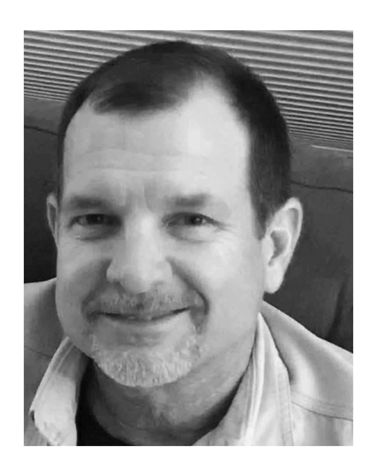 Black and white close-up photo of smiling middle-aged man with short dark hair, light facial hair, wearing a light-colored shirt, in an indoor setting.